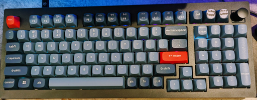 A close-up view of a dark grey Keychron V5 Max mechanical keyboard featuring grey and blue keycaps and a prominent red Esc key and red Enter key. The top right corner includes a programmable rotary encoder knob. The keyboard is backlit. A temporary piece of tape with handwritten text is stuck to the top right keycaps, saying "Print Screen" and "Scroll Lock"